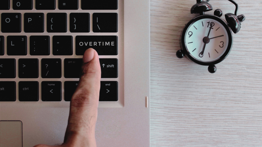 A clock showing 6 o'clock and a hand reaching for an "overtime" button on a laptop keyboard.